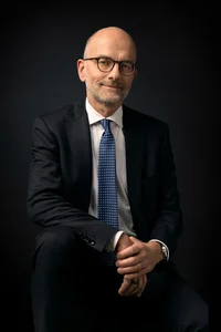 Sitzender Mann mit Brille, dunklem Anzug und blauer Krawatte