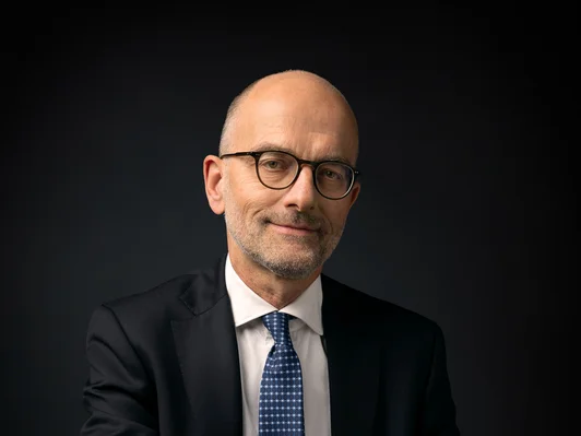 Sitzender Mann mit Brille, dunklem Anzug und blauer Krawatte