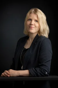 Frau mit schulterlangem, blonden Haar. An einem Tisch stehend, den Arm darauf abgestützt.