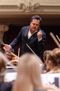 Dirigent Robert Trevino in ausdrucksvoller Geste: Mit geschlossenen Augen beugt er sich dem Orchester zu, eine Hand zur Faust geballt, die andere weit geöffnet. Im Vordergrund sind unscharf Streicher mit erhobenen Bögen zu sehen, im Hintergrund eine verzierte Stuckdecke.