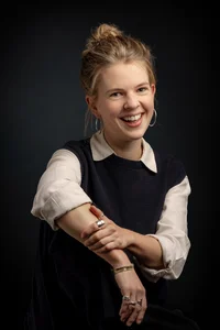 Frau mit blonden, zurückgebundenen Haaren. Den Arm auf einem Knie aufgestützt.