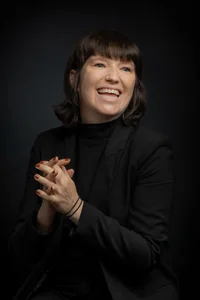 Frau mit braunen, schulterlangen Haaren. Schwarzer Blazer, die Hände gefaltet.