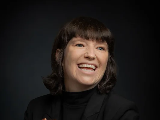 Frau mit braunen, schulterlangen Haaren. Schwarzer Blazer, die Hände gefaltet.
