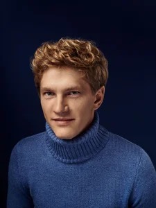 Porträt des Pianisten Jan Lisiecki vor dunkelblauem Hintergrund, blondes Haar, blauer Rollkragenpullover.