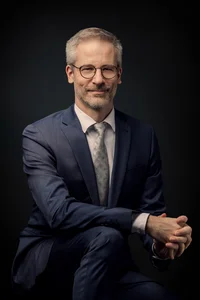 Mann, mit kurzem grau-meliertem Haar und Brille. Dunkelblauer Anzug, sitzend.