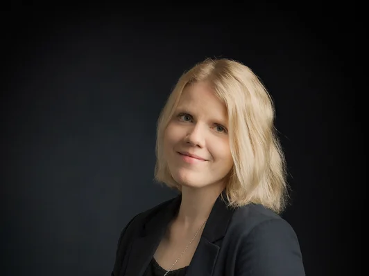 Frau mit schulterlangem, blonden Haar. Aufrecht sitzend mit schwarzem Blazer.