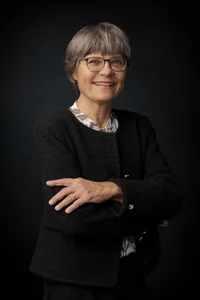 Frau mit kurzem, grauen Haar und Brille und schwarzem Blazer. Stehend, die Arme überschränkt.