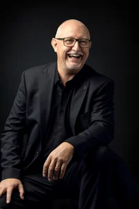 Mann mit Brille und Kinnbart in schwarzem Anzug. Sitzend und lachend.
