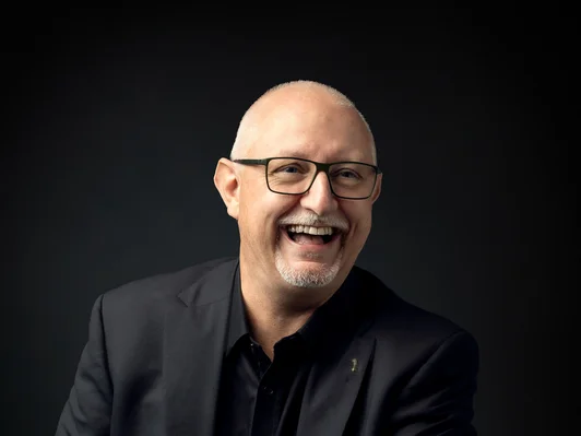 Mann mit Brille und Kinnbart in schwarzem Anzug. Sitzend und lachend.