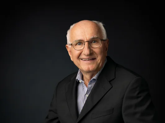 Mann mit weissem Haar und Brille. Schwarzer Anzug, sitzend.