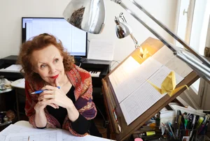 Die Komponistin Kaija Saariaho an ihrem Schreibtisch, das Kinn auf die Hände gestützt, mit Kugelschreiber, Partitur und Origami-Kranichen auf dem Notenpult.