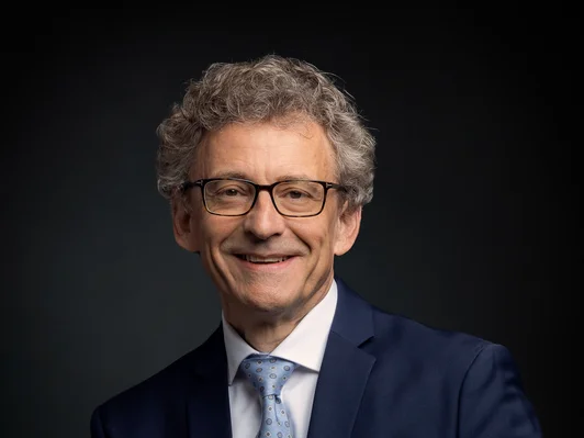 Mann, sitzend, mit Brille und blauem Anzug.
