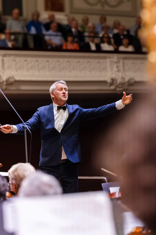 Markus Poschner dirigiert das Sinfonieorchester Basel, Taktstock in der ausgestreckten Hand, dunkelblauer Anzug mit Fliege.