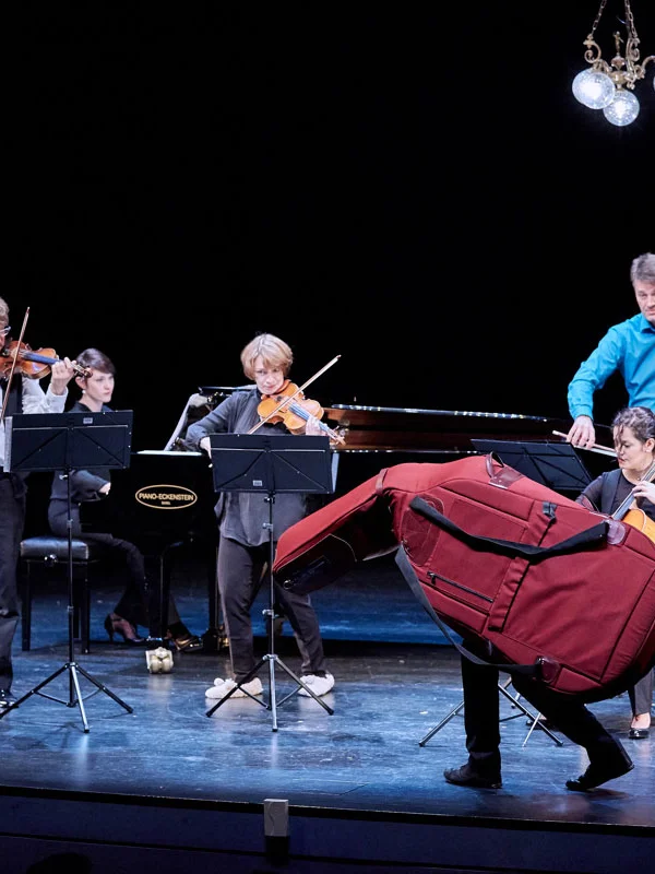 Bühne mit Musiker*innen, ein Musiker versteckt sich in einer Cello-Hülle