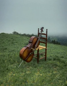 Ein Cello mit Bogen lehnt an einem Holzstuhl mit geflochtener Sitzfläche. Der Stuhl steht allein auf einer grünen Anhöhe, im Hintergrund liegt Nebel über der Landschaft.